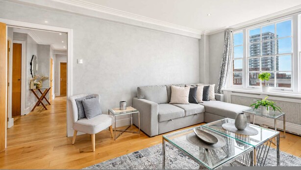 A modern living room with a grey sofa, a glass coffee table, and a view of a cityscape through large windows. - Beautiful 2 Bedroom off Marble Arch (London)
