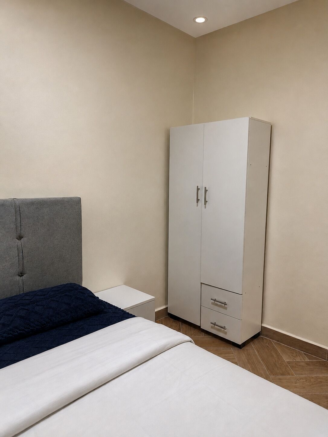 A bedroom with a bed, a grey headboard, a white wardrobe with drawers, and a small bedside table.