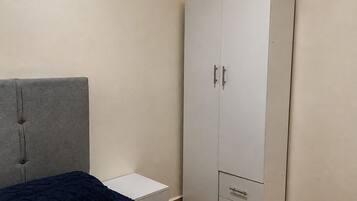 A bedroom with a bed, a grey headboard, a white wardrobe with drawers, and a small bedside table.
