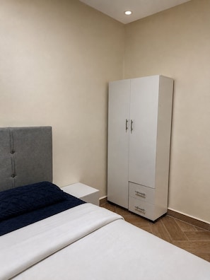 A bedroom with a bed, a grey headboard, a white wardrobe with drawers, and a small bedside table.