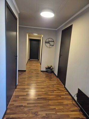 Um corredor com piso de madeira, luminária no teto, relógio na parede e um vaso com planta.