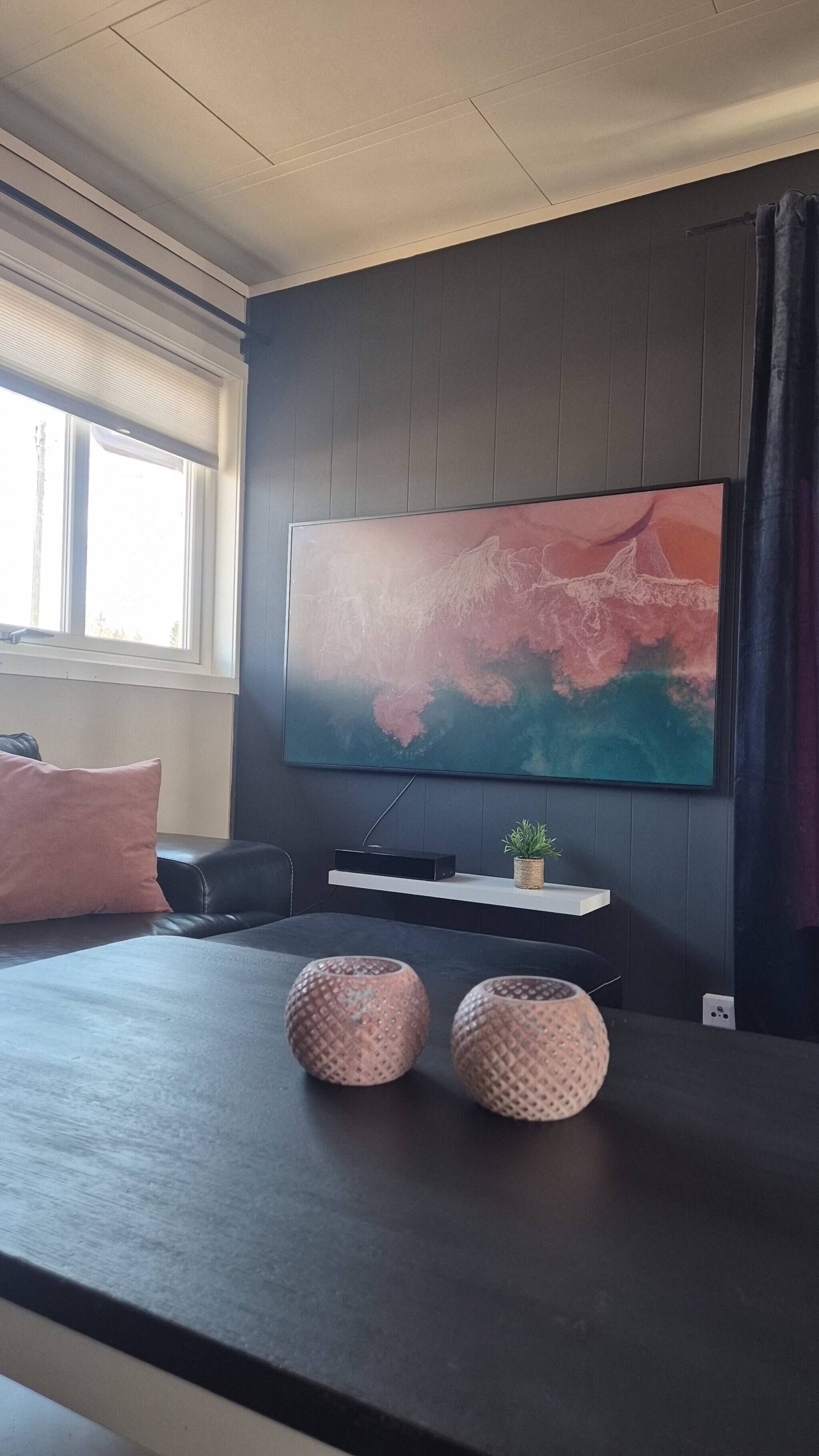 A modern living room with a large flat-screen TV mounted on the wall, a dark wooden coffee table, two decorative vases, a small plant, and a black leather sofa with a pink pillow.