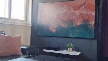 Una sala moderna con un televisor de pantalla plana grande montado en la pared, una mesa de centro de madera oscura, dos jarrones decorativos, una planta pequeña y un sofá de cuero negro con un cojín rosa.