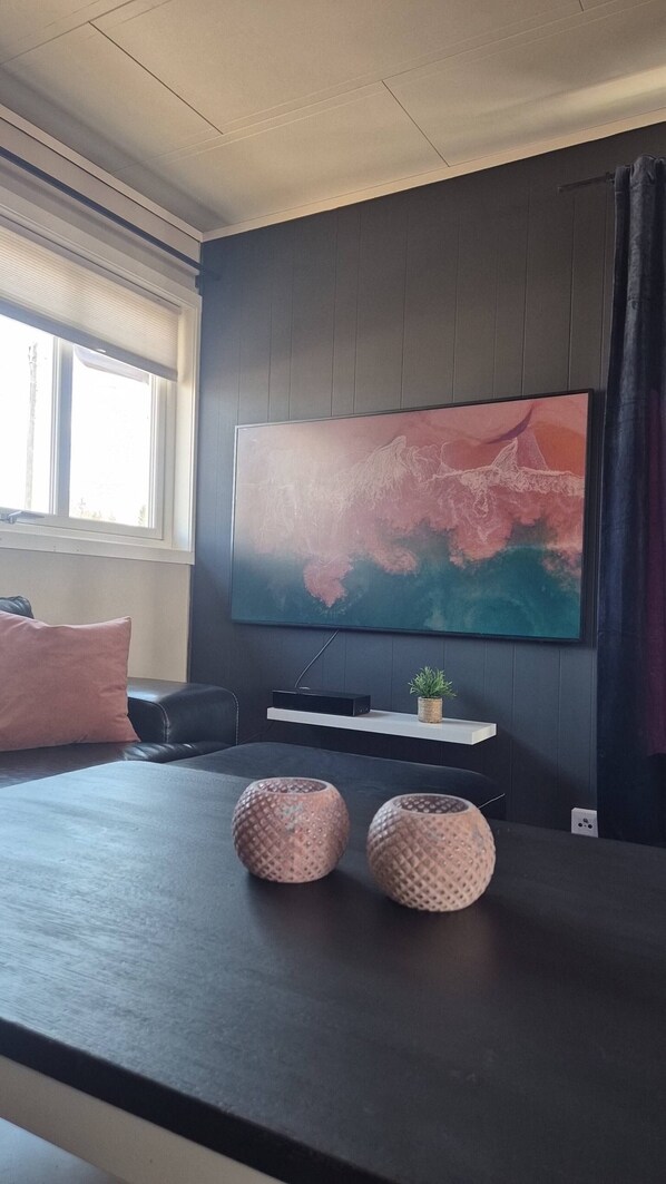 A modern living room with a large flat-screen TV mounted on the wall, a dark wooden coffee table, two decorative vases, a small plant, and a black leather sofa with a pink pillow.