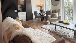 A modern living room with a brown sofa, a dining area with a table and chairs, a kitchen in the background, and a large window providing natural light. - Executive 2-bedroom Corporate Space In West Yorkshire, United Kingdom (Leeds)