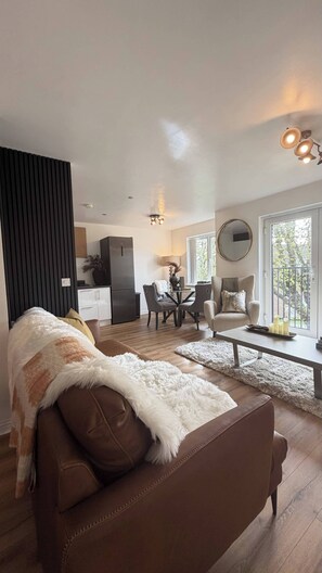 Ein modernes Wohnzimmer mit einem braunen Sofa, einem Essbereich mit Tisch und Stühlen, einer Küche im Hintergrund und einem großen Fenster, das natürliches Licht hereinlässt.