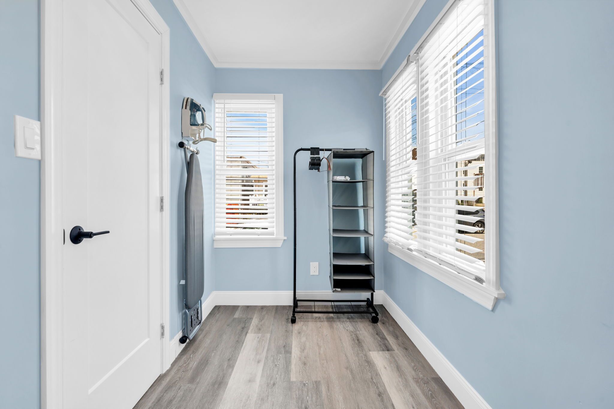 Un couloir aux murs bleu clair, une porte blanche et un plancher en bois. On y trouve deux fenêtres avec des stores blancs, un présentoir en métal noir doté d’étagères et un plafond blanc.
