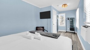 A hotel room with a bed, a flat-screen TV, a chair, a window with blinds, and a wall-mounted heater.