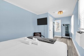 A hotel room with a bed, a flat-screen TV, a chair, a window with blinds, and a wall-mounted heater. - King Bed | Half Block to Beach | Prof Managed (Seaside Heights)