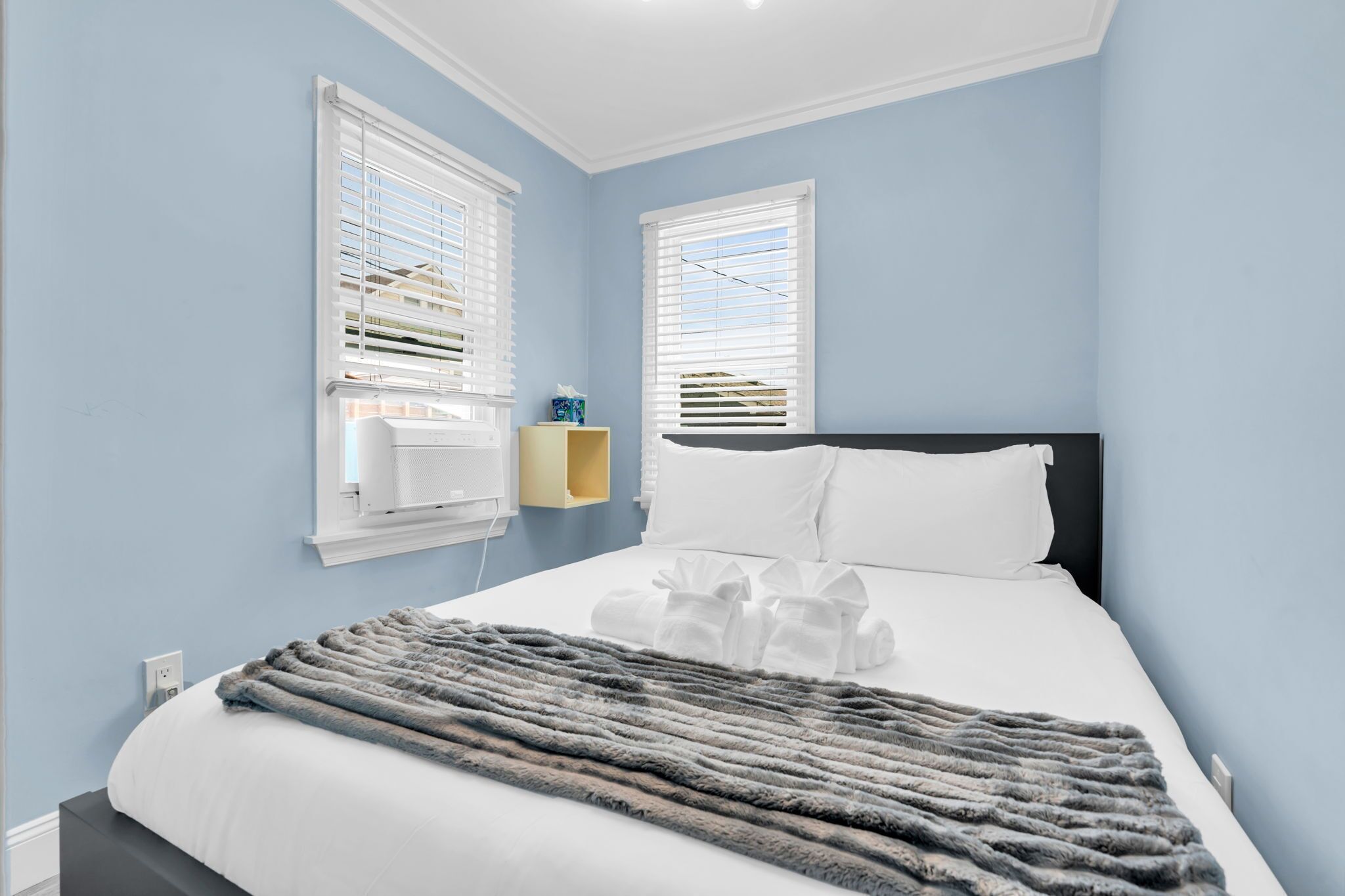 A bedroom with a bed, two windows with blinds, a bedside table with a tissue box, and a grey blanket.