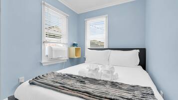 A bedroom with a bed, two windows with blinds, a bedside table with a tissue box, and a grey blanket.