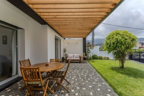 Un patio exterior con una mesa y sillas de madera, un sofá con cojines y una pequeña zona ajardinada con un árbol.