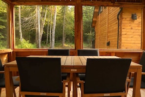 A wooden cabin with a dining table and chairs, large windows, and a view of a forest. - Sauna, Spa & Nature | Le Kanouk (Lac-Etchemin)