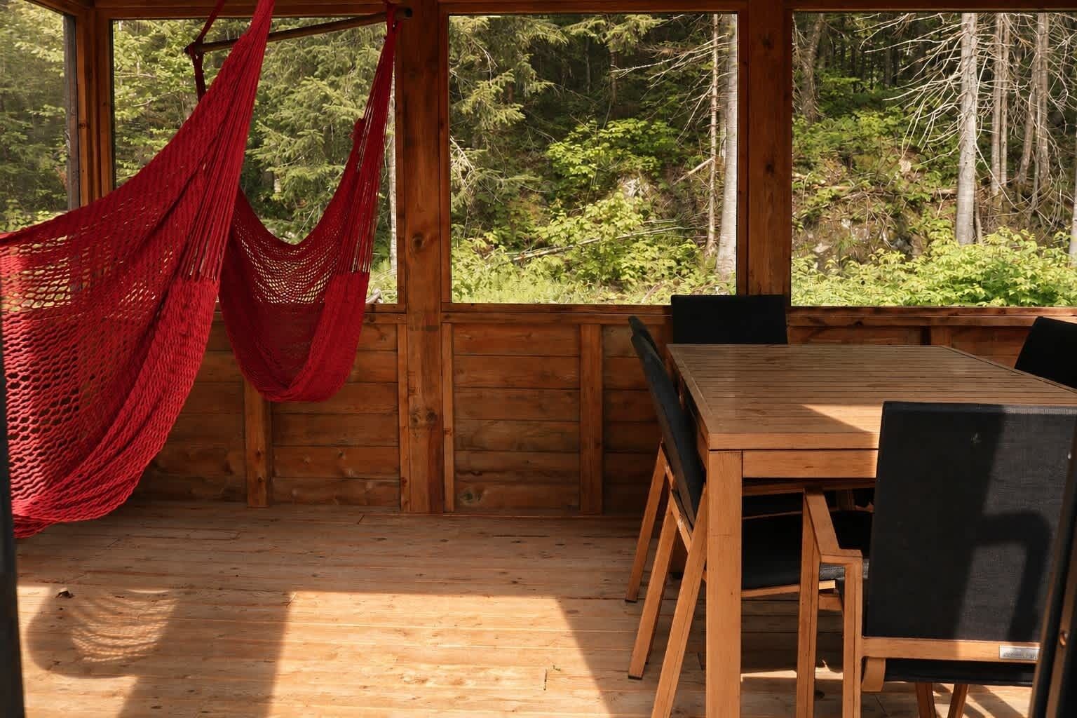 Une terrasse en bois avec une table et des chaises, un hamac rouge et une vue sur une forêt.