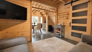A cozy cabin interior with a TV, a dining area, and a staircase leading to an upper level. - Sauna, Spa & Nature | Le Kanouk (Lac-Etchemin)