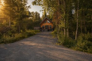 A cabin with a gravel driveway leading to a wooden door, surrounded by dense forest. - Sauna, Spa & Nature | Le Kanouk (Lac-Etchemin)