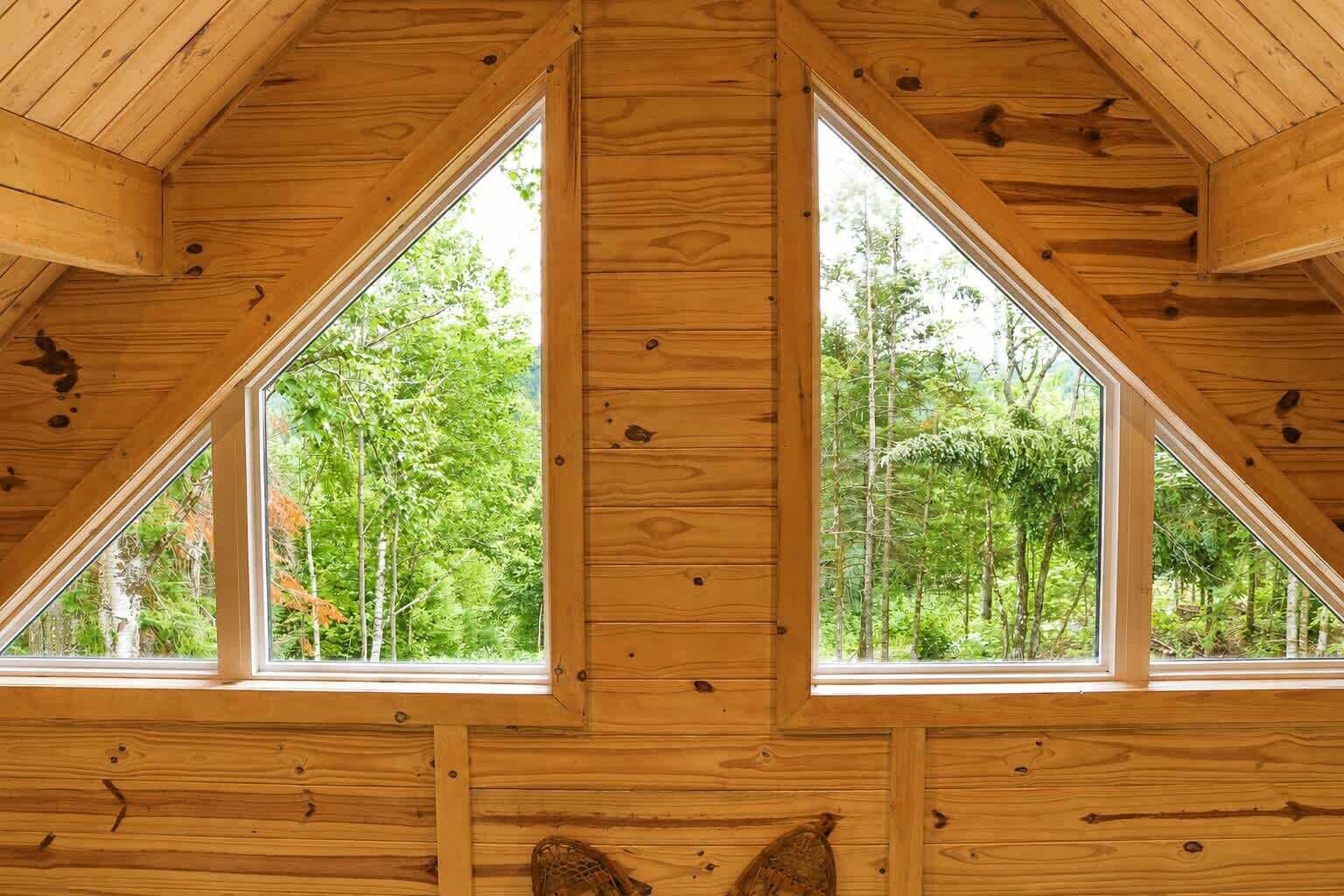 A wooden cabin interior with a sloped roof and large windows offering a view of a forest.