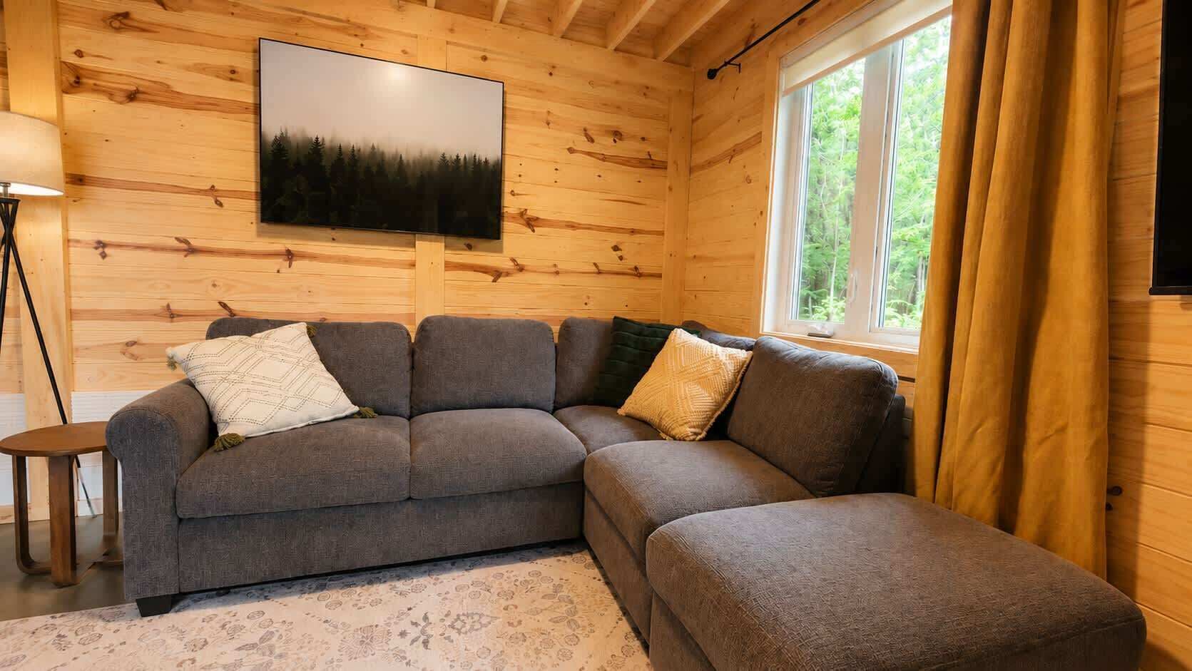 Intérieur moderne d’une cabane en rondins, avec un canapé d’angle, un téléviseur à écran plat et une fenêtre avec des rideaux.