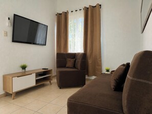 Ein Wohnzimmer mit flachbildfernseher an der Wand, einem braunen Sofa, einem Holznachttisch mit einer Pflanze und einem Fenster mit Vorhängen.