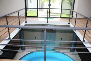 An indoor swimming pool with a glass barrier, wooden flooring, and a view of a green outdoor area. - The Glass & Stones Oasis: An Urban Sanctuary (Temixco)