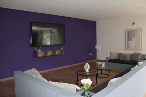 Una sala de estar moderna con un televisor de pantalla plana montado en una pared morada, un sofá gris, una mesa de centro y una lámpara de pie.