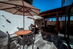 An outdoor patio with wooden tables and chairs, umbrellas, and a brick wall. - Apto com Piscina Academia e Churrasqueira (João Pessoa)