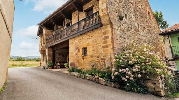 A stone building with a balcony, a paved road in front, and a flower bed with pink flowers. - Casa Los Josuco, located in the heart of the Saja-Besaya Natural Park. (Collado de Cieza)