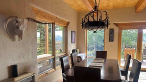A dining area with a wooden table and chairs, large windows with a mountain view, a chandelier, and a mounted animal head on the wall. - Studley Ranch-Private hot tub and pet friendly (Crested Butte)