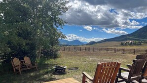 A wooden deck with a table and chairs, a fire pit, and a view of a fenced field and mountains. - Studley Ranch-Private hot tub and pet friendly (Crested Butte)