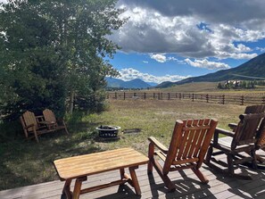 Eine Holzterrasse mit Tisch und Stühlen, einer Feuerstelle und Blick auf ein eingezäuntes Feld und Berge.