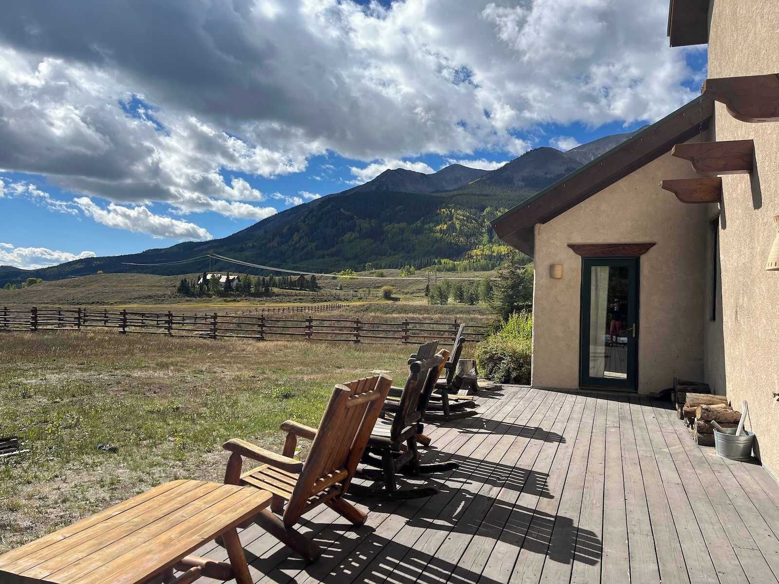 A wooden deck with chairs and tables, a building with a door, and a mountainous landscape in the background.