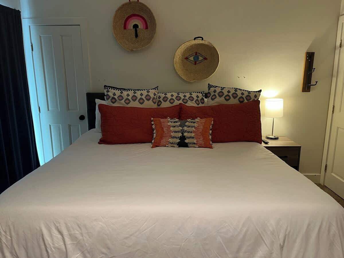 A bed with white bedding, red and patterned pillows, two wall-mounted hats, a bedside lamp, and a door.