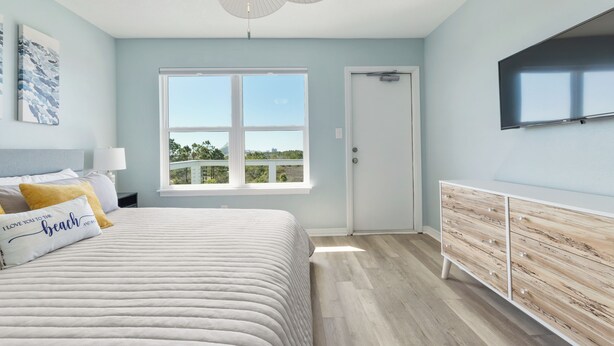 Um quarto com cama, mesa de cabeceira, cômoda, ventoinha no teto, janela e televisão fixada na parede.