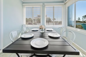 A dining table set for four with white chairs, a vase with flowers, and a view of buildings through the window blinds. - Luxury Beach House, Heated Pool & Spa, Sauna, Game Room, Private Beach Access (Pensacola)