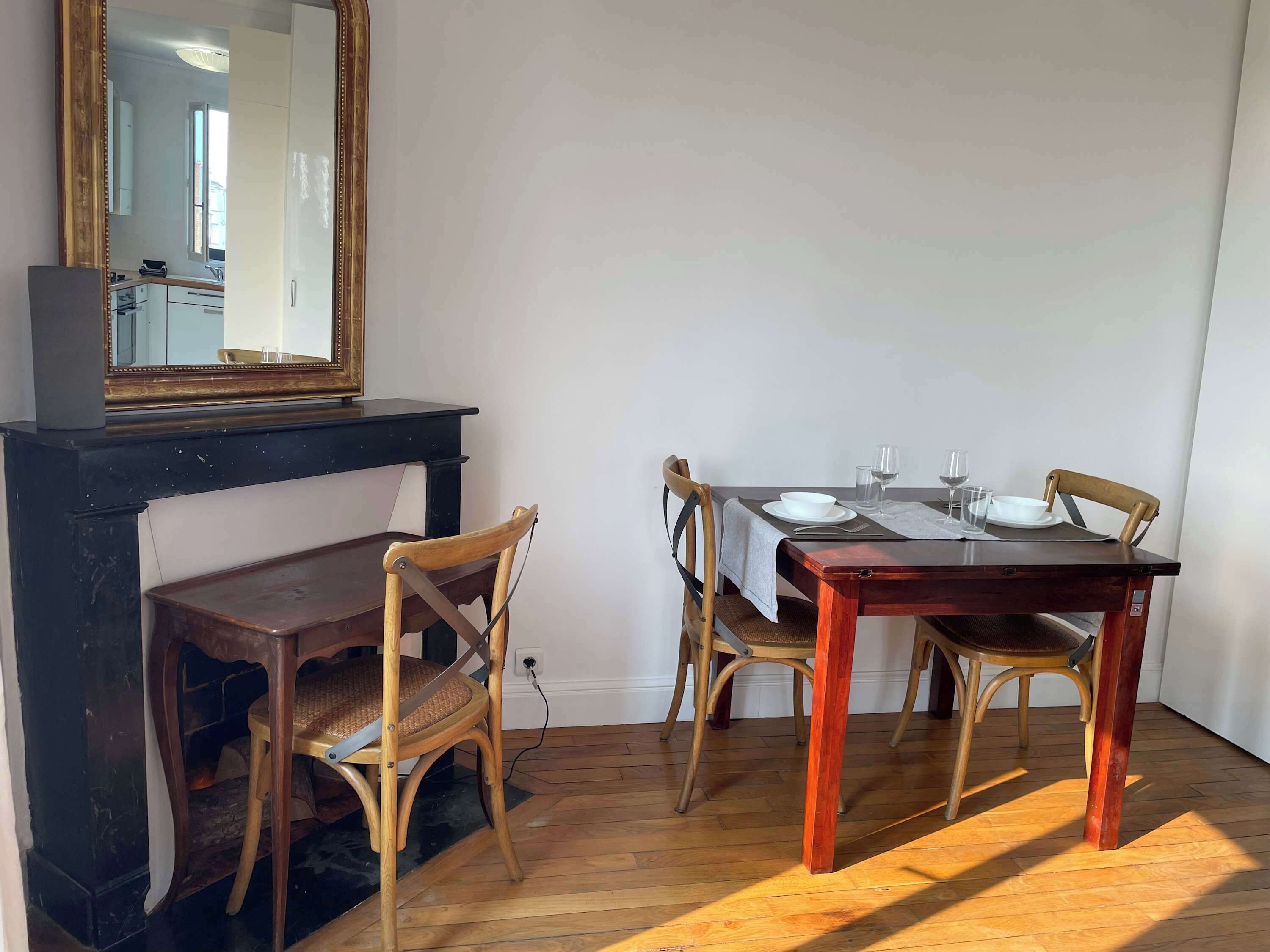 A dining area with a wooden table, four chairs, a mirror above the table, and a fireplace below.