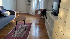 Ein modernes Wohnzimmer mit einer Couch, einem kleinen Tisch, einem Korbstuhl, einem Fernseher und Blick auf die Stadt durch große Fenster.