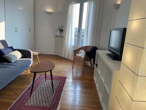 Ein modernes Wohnzimmer mit einer Couch, einem kleinen Tisch, einem Korbstuhl, einem Fernseher und Blick auf die Stadt durch große Fenster.