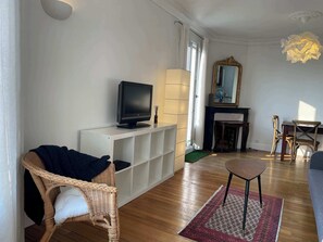 Ein Wohnzimmer mit einem Fernseher auf einem weißen Schrank, einem Korbstuhl, einem kleinen Tisch und einem gemusterten Teppich.