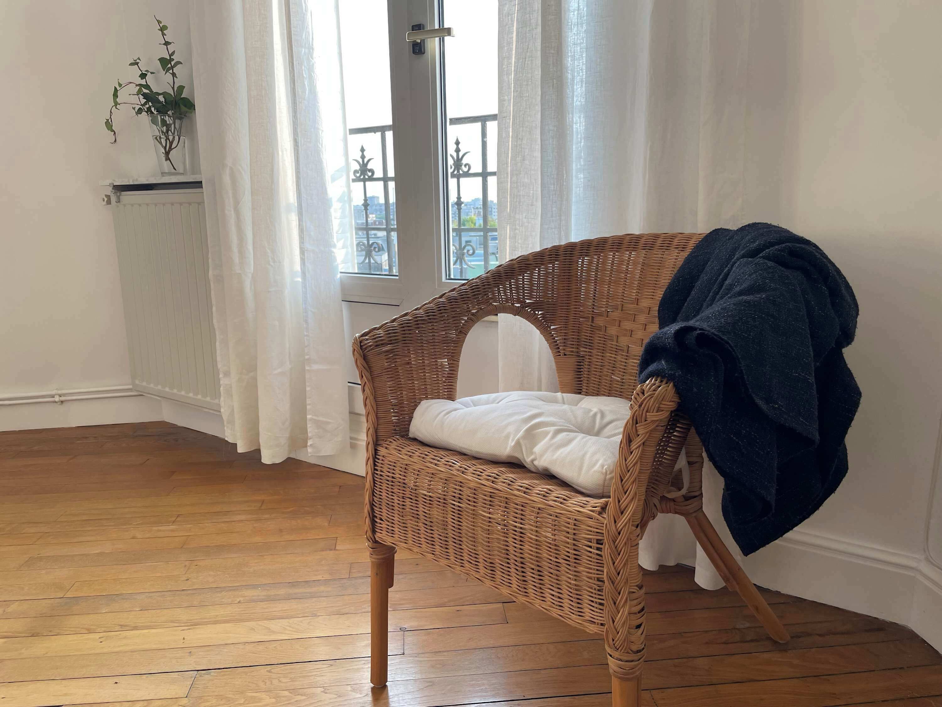Una silla de mimbre con un cojín y una manta, ubicada cerca de una ventana con cortinas blancas.