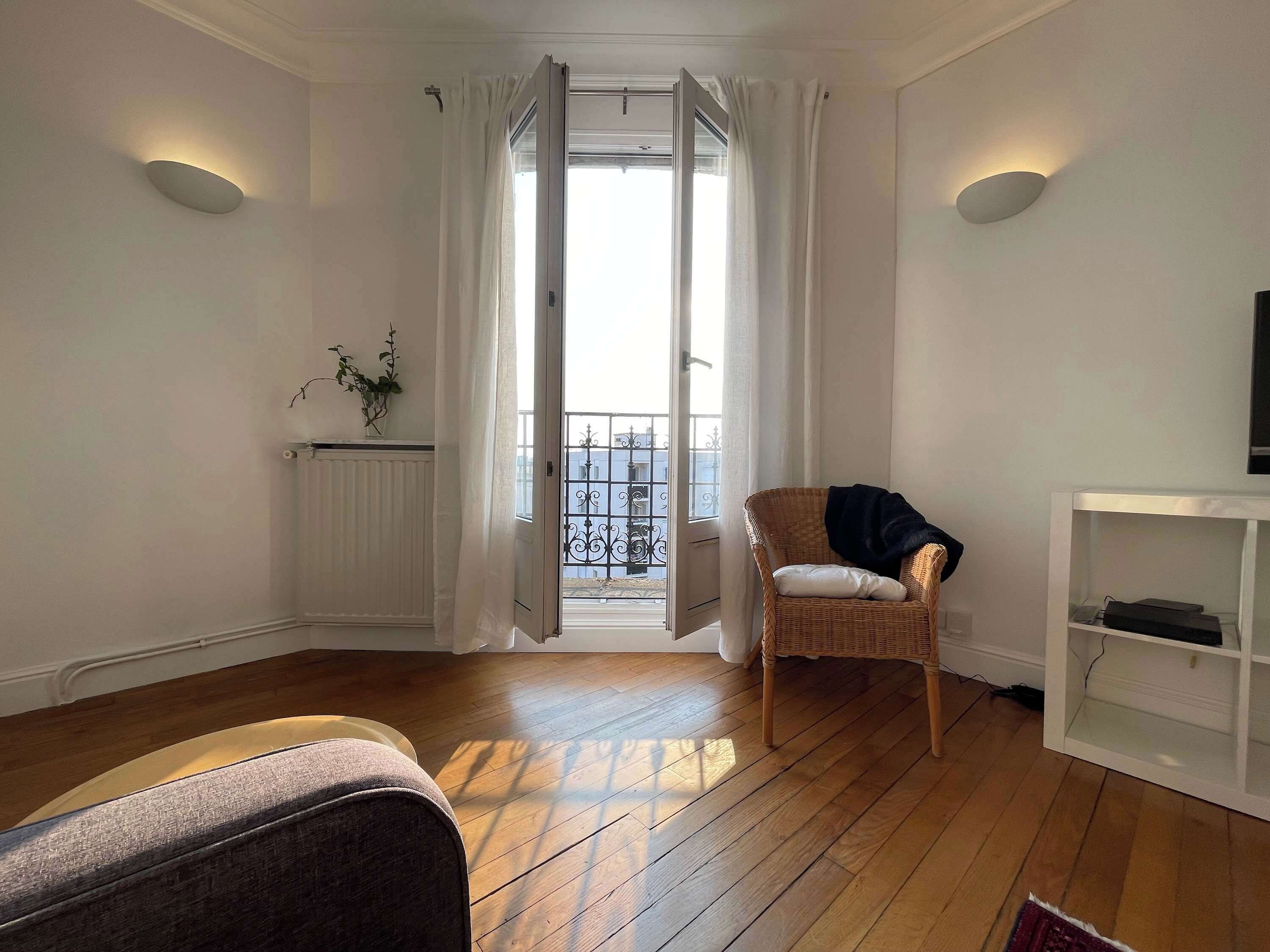 Une pièce avec un plancher en bois, une chaise en osier, un radiateur blanc, une petite plante et une console blanche sur laquelle se trouve un téléviseur.