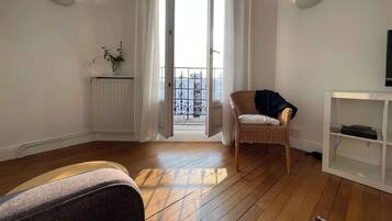 Une pièce avec un plancher en bois, une chaise en osier, un radiateur blanc, une petite plante et une console blanche sur laquelle se trouve un téléviseur.