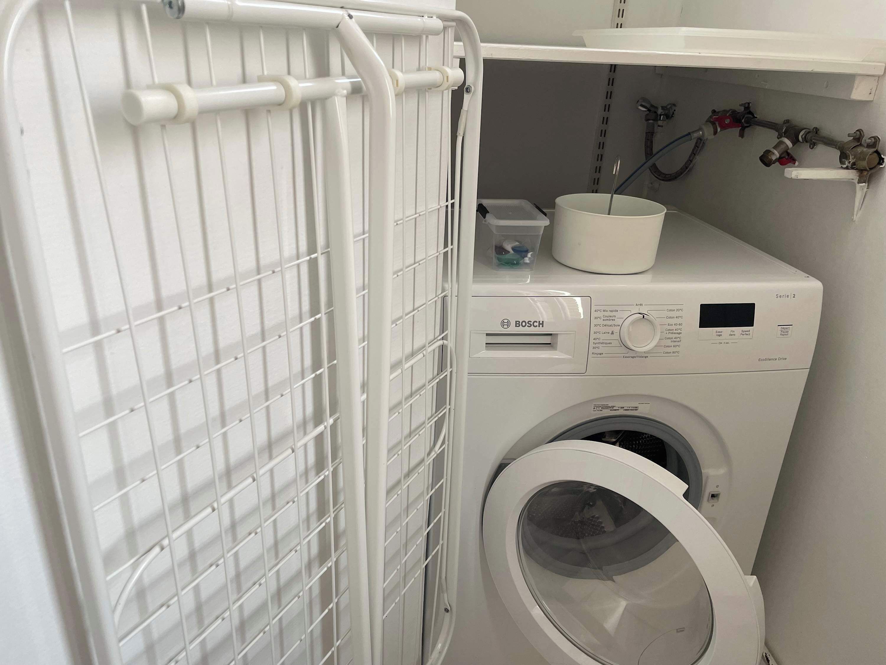 A compact, white Bosch washing machine with a control panel and a front-loading door, situated in a small laundry area with a metal drying rack and a plastic container on top.