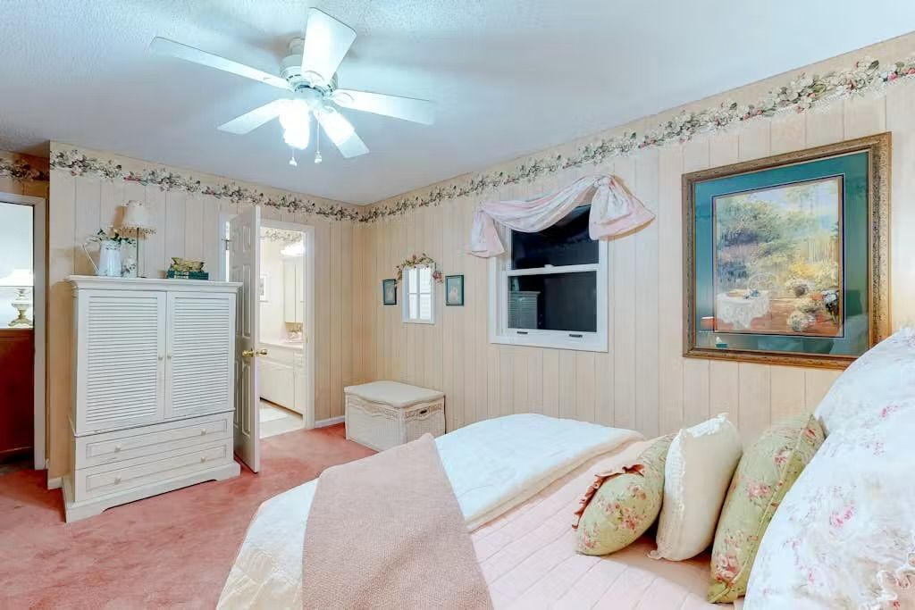 Une chambre à coucher avec un lit, une armoire, une petite table, une fenêtre et un ventilateur de plafond.