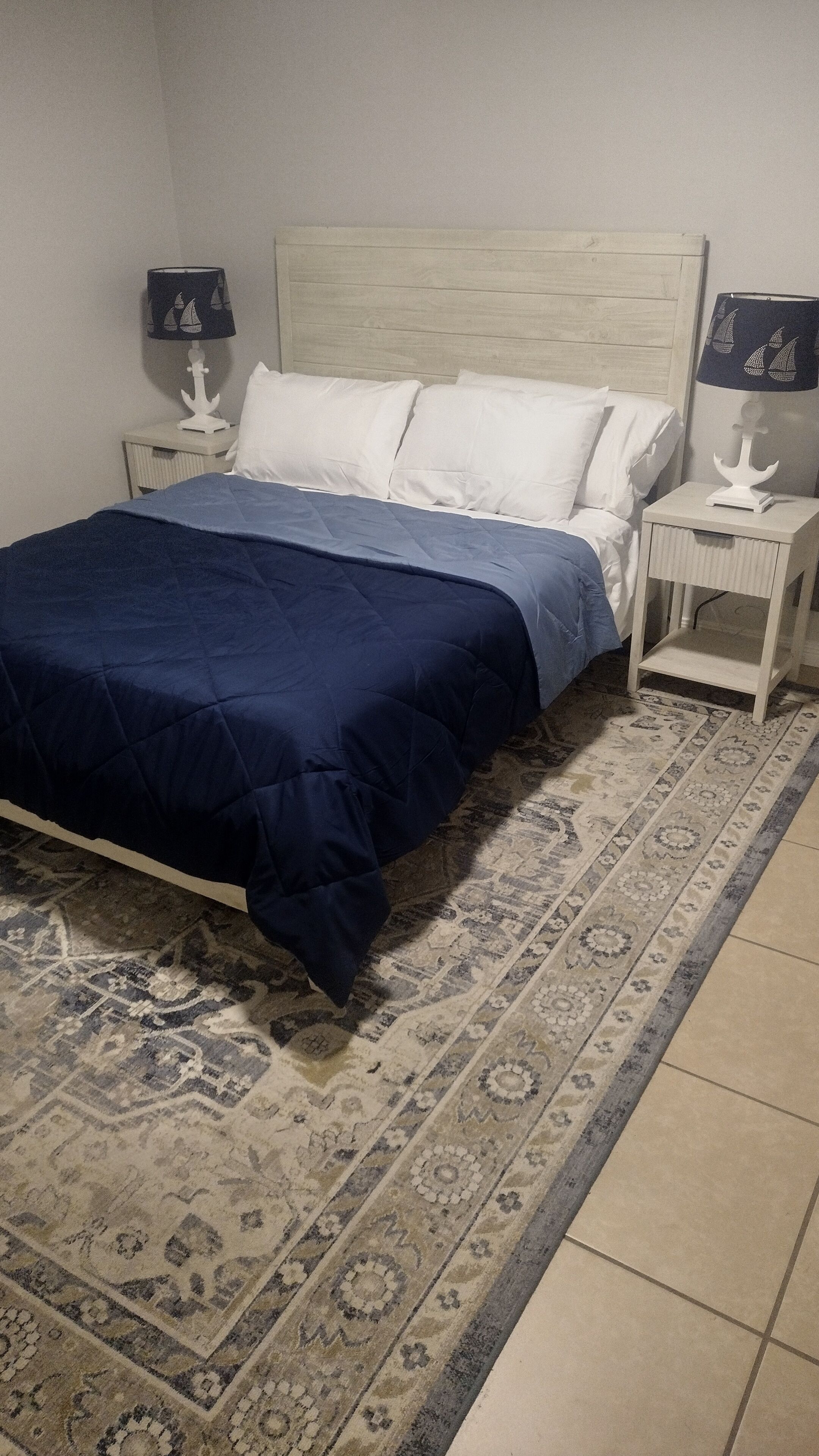 A bed with a blue bedspread, white pillows, and a headboard. There are two bedside tables with lamps, and a patterned rug on the floor.