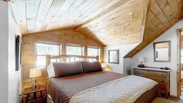 A bedroom with a wooden ceiling, a bed with a quilted cover, a bedside table with a lamp, and a dresser. - The Dragonfly  Studio (South Fork)