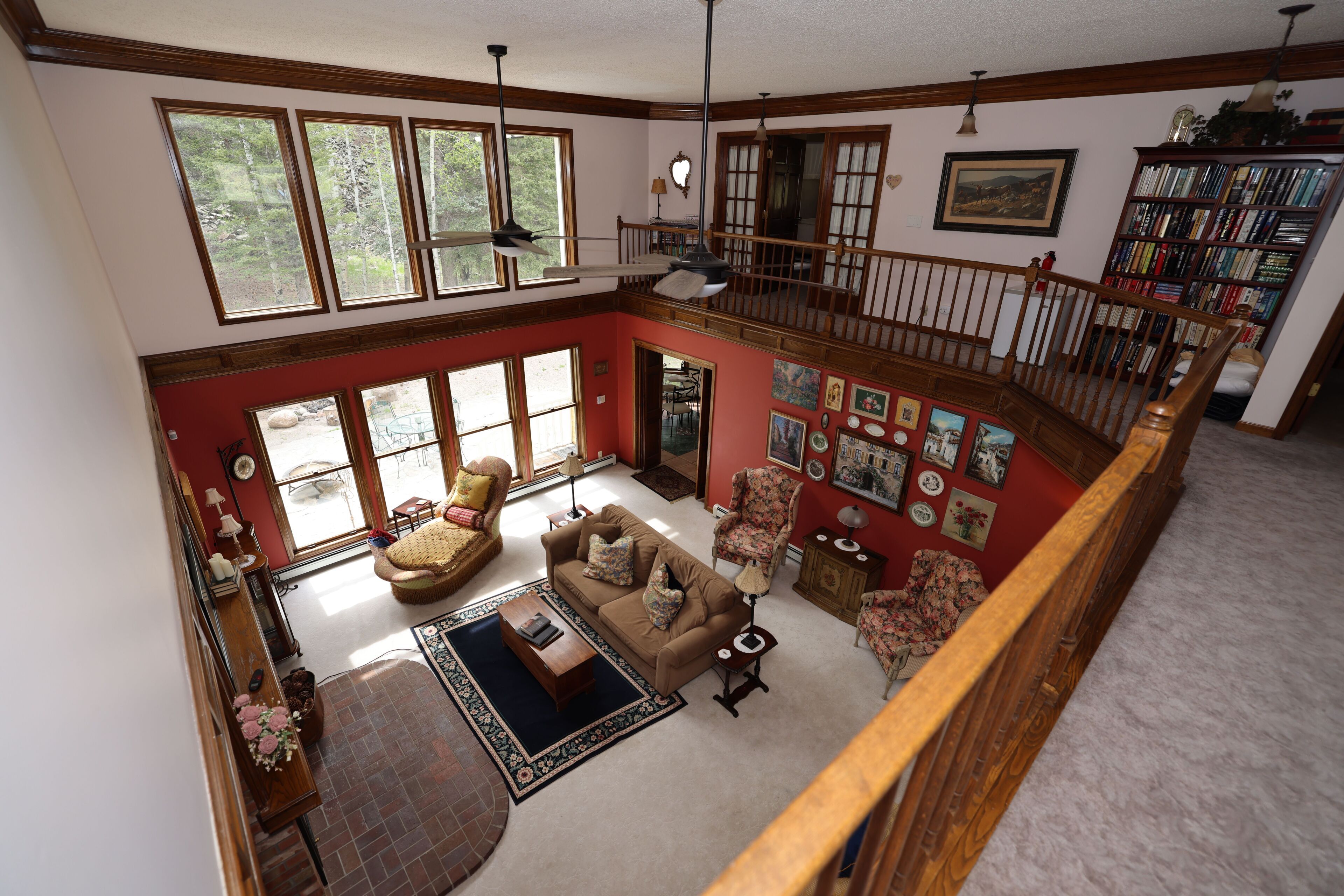 A spacious living area with a high ceiling, large windows, a wooden staircase, and a bookshelf.