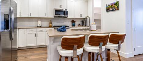 A modern kitchen with white cabinets, a stainless steel refrigerator, a microwave, and a gas stove. There are three wooden bar stools with metal legs.
