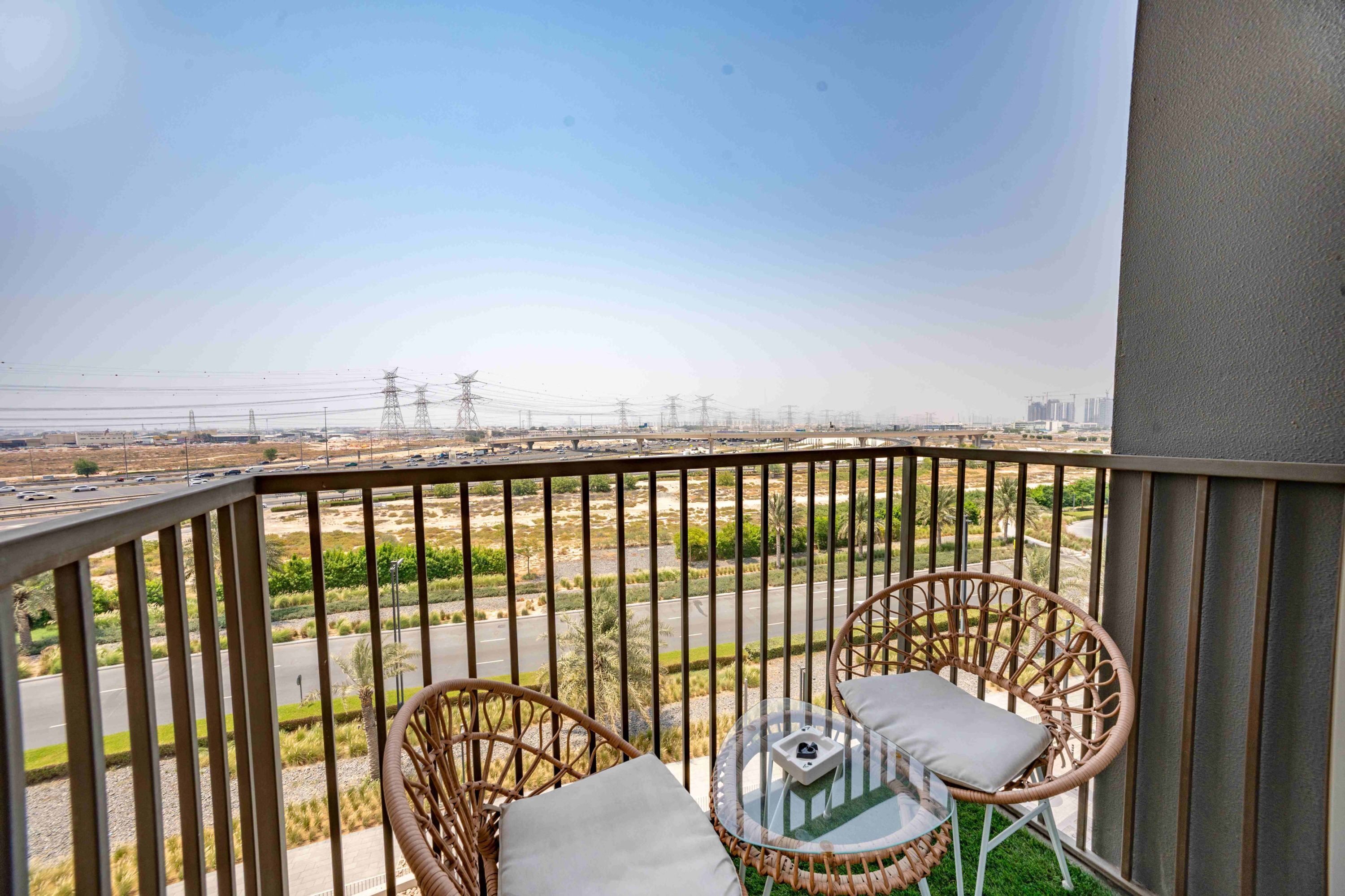 A balcony with wicker chairs and a small table, overlooking a road and distant buildings.