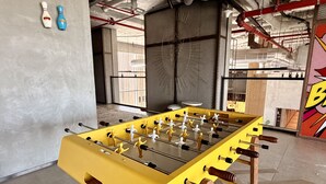 Uma mesa de futebol em uma sala com piso de concreto, vigas de metal e uma grande janela.