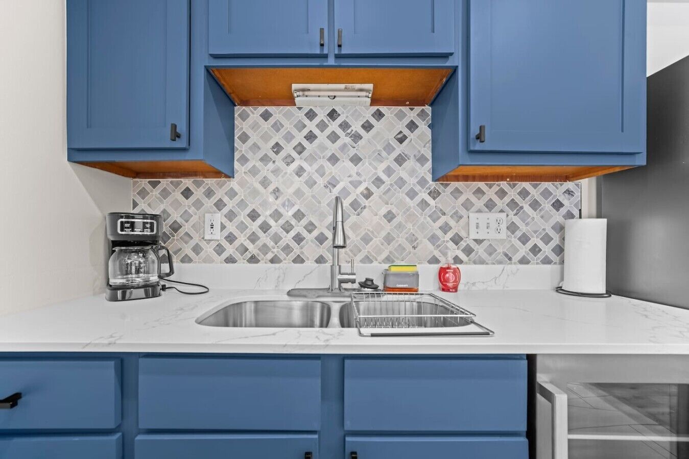 A kitchen with blue cabinets, a white countertop, a coffee maker, and a tiled backsplash.